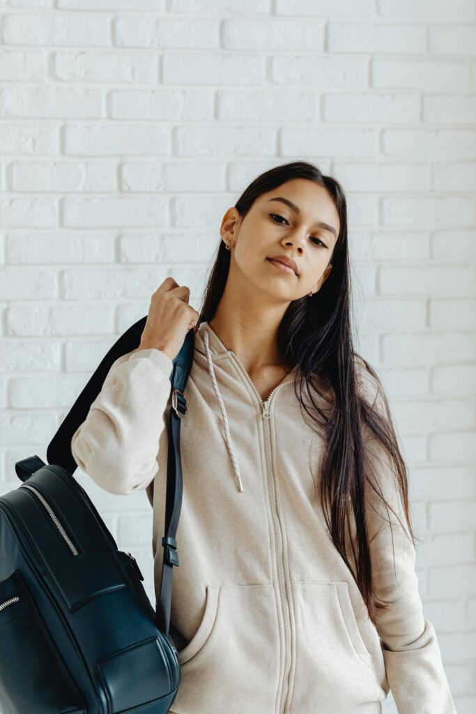 Confident teenager with a backpack and hoodie poses against a white brick wall.