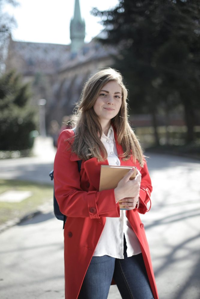 pexels-photo-3793238-3793238 Young woman in a red coat smiling while holding a book outside on a sunny day.